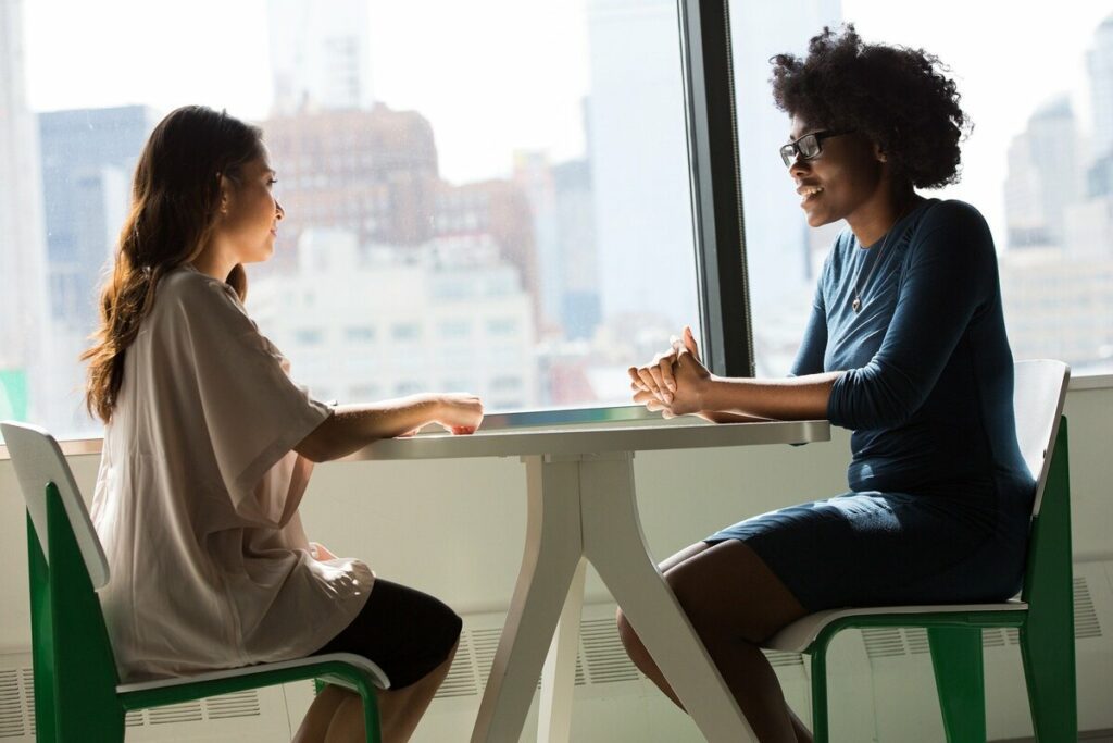 Zwei Frauen in Businesskleidung unterhalten sich an einem Tisch vor einer Glasfenster-Front