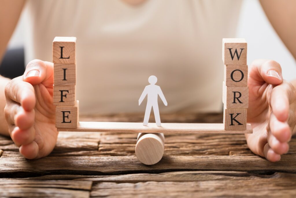 Holzwippe mit einem Holzmännchen, auf der linken Seite Würfel mit schwarzen Buchstaben LIFE auf der anderen Seite WORK
