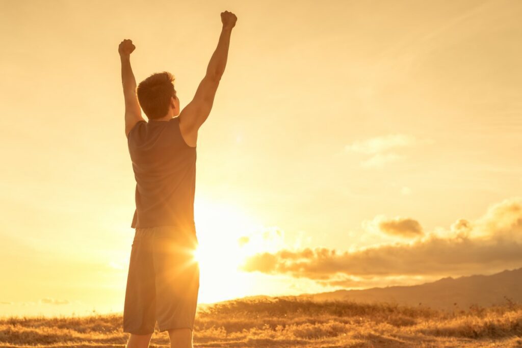 Mann steht auf einem Feld bei Sonnenaufgang und streckt in Siegerpose beide Arme in die Luft