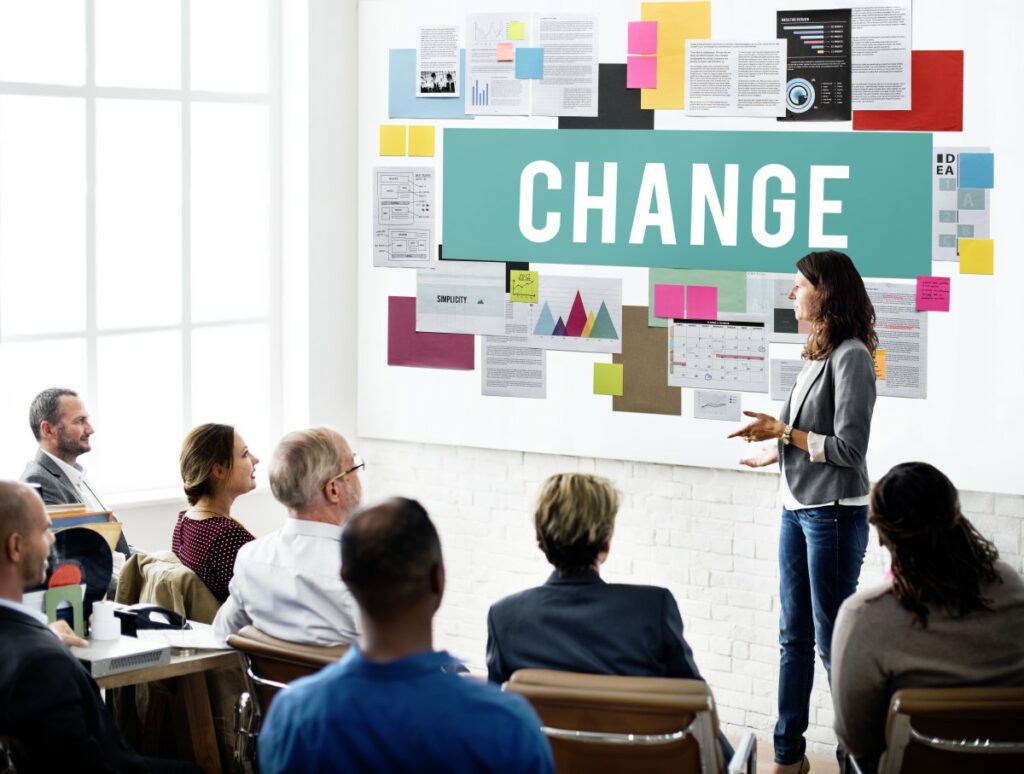 Frau in Jeans mit grauem Blazer steht vor einer Gruppe Erwachsener und erklärt - am Whiteboard hinter ihr groß das Wort CHANGE und verschiedene Dokumente und Charts