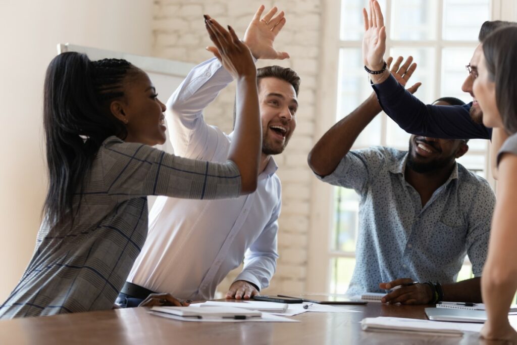 Fünf Mitarbeiter und Mitarbeiterinnen sitzen um einen Besprechungstisch und geben sich ein High Five
