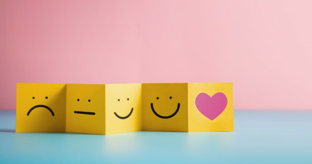 Gelbes Feedback Leporello mit verschiedenen Smileys und einem rosa Herz auf rosa-blauem Hintergrund