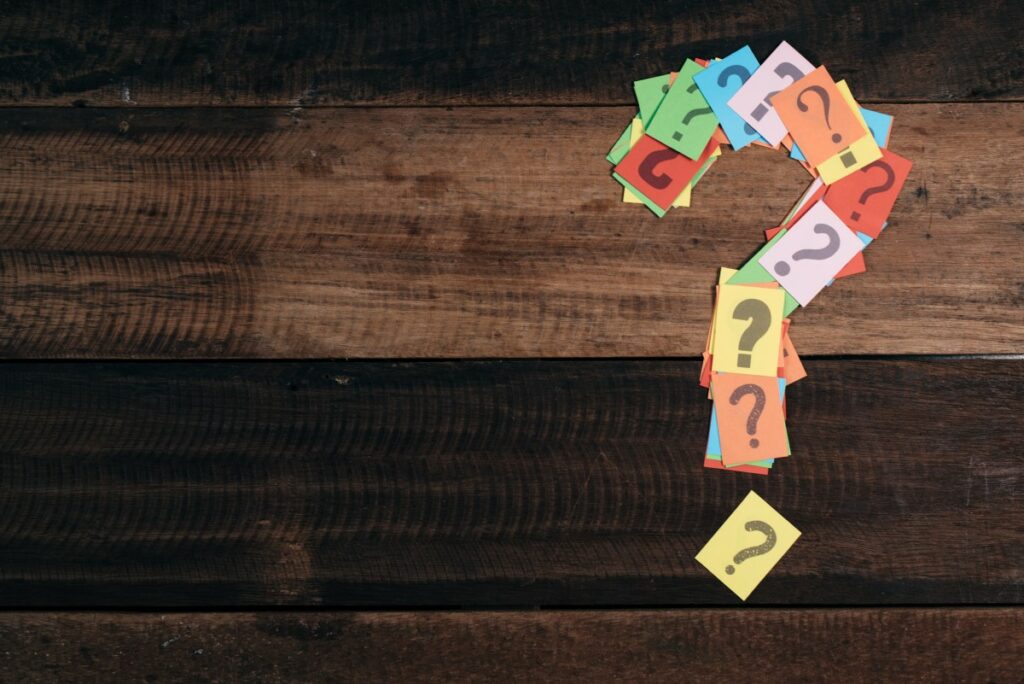 Bunte Post-Its mit schwarzen Fragezeichen, angeordnet als Fragezeichen auf einem Holztisch