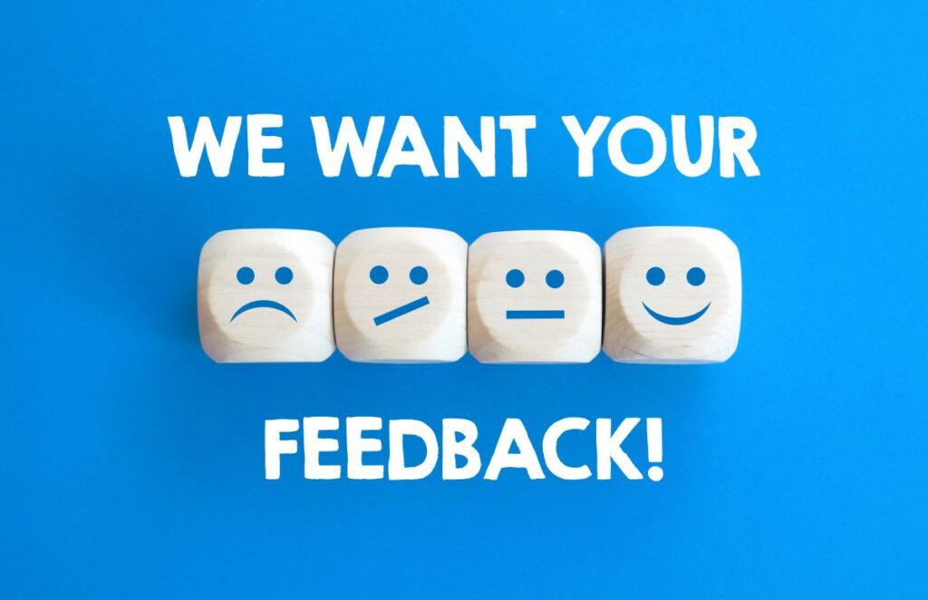 Vier weiße Würfel vor blauem Hintergrund mit aufgemalten Gesichtern von traurig bis glücklich - in weißer Schrift we want your feedback
