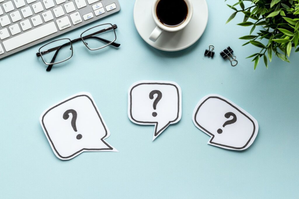 Hellblauer Schreibtisch mit Kaffeetasse, Brille und drei Sprechblasen in weiß mit schwarzen Fragezeichen