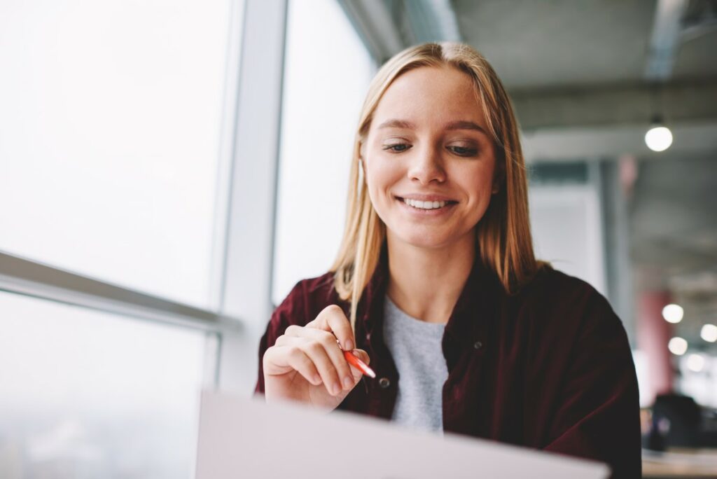 Blonde Frau sitzt am Schreibtisch mit Stift in der Hand und lächelt